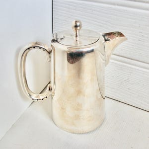 Op de afbeelding: Een zilverkleurige koffiepot met een gebogen handvat en een deksel met een kleine knop. De pot heeft een tuit om uit te schenken en is gemaakt van glanzend, reflecterend metaal. Het ontwerp is eenvoudig en elegant.