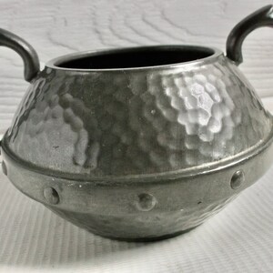 May include: A hammered pewter sugar bowl with two handles and a textured surface. The bowl has a rounded shape and a band of decorative studs around the rim.