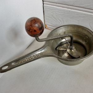May include: A vintage silver food mill with a wooden handle. The mill has a strainer and a crank. The handle is labelled "Moulie-Baby".
