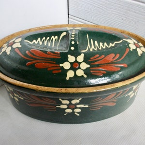 May include: A green oval ceramic casserole dish with a lid. The dish is decorated with white flowers and red leaves. The lid has a small hole in the center.