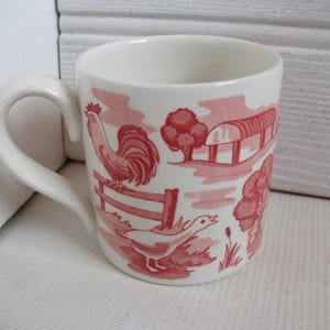 Puede incluir: Taza de cerámica blanca con un diseño de escena de granja en rojo. El diseño incluye un gallo, un ganso, una valla, árboles y un granero. La taza tiene un asa curva.