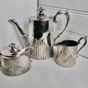Op de afbeelding: Een zilveren theeservies met een theepot, roomkan en suikerpot. Elk stuk heeft een decoratief, verticaal, geribbeld ontwerp.