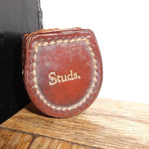 Puede incluir: Una funda de cuero marr&oacute;n con la palabra "Studs." grabada en letras doradas.