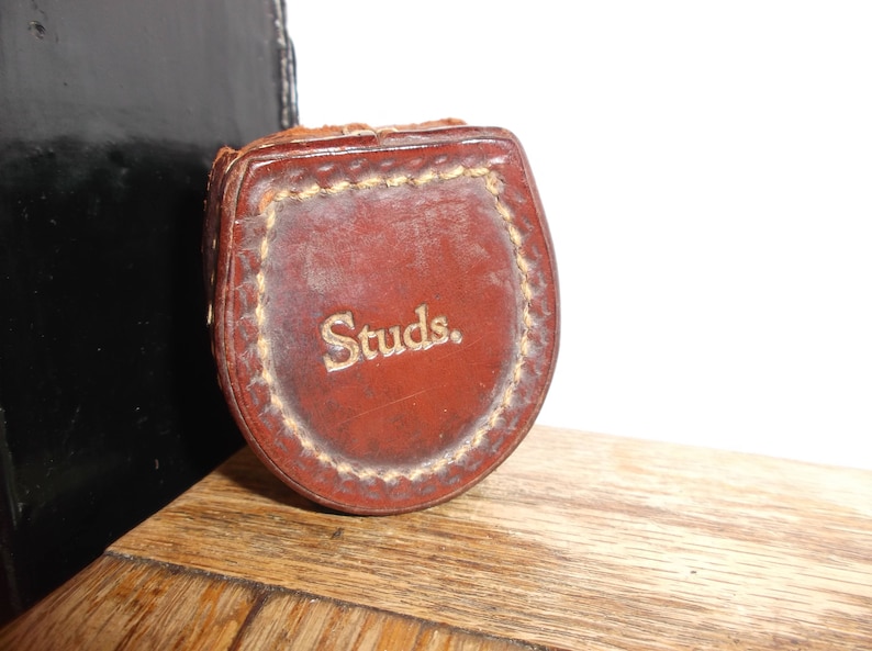 Puede incluir: Una funda de cuero marr&oacute;n con la palabra "Studs." grabada en letras doradas.