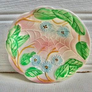 Op de afbeelding: Een klein, wit keramiek schaaltje met een roze en groen bloemenontwerp. Het schaaltje heeft een spinnenwebontwerp in het midden en is versierd met blauwe bloemen en groene bladeren.