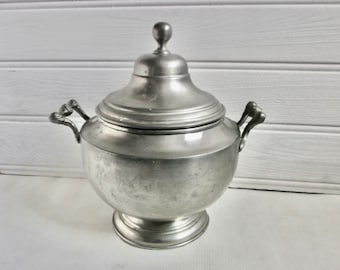 May include: A silver pewter serving dish with a lid and two handles. The dish has a round body and a raised base.