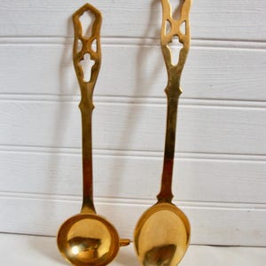 May include: Two vintage brass serving spoons. The spoons have a long, ornate handle with a decorative cutout design. The spoon bowls are round and polished. The spoons are displayed against a white, horizontal-slat background.