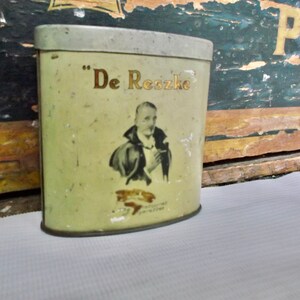 Puede incluir: Una lata de metal vintage con fondo crema y letras doradas. La lata presenta un retrato de un hombre con traje oscuro y camisa blanca con corbata negra. El texto en la lata dice "De Reszke".