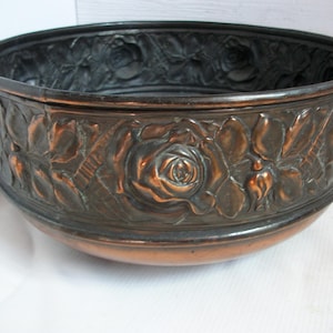 Könnte beinhalten: Eine kupferfarbene Metallschale mit dunklem Innenraum und floralem Rosendesign. Die Außenseite der Schale weist geprägte Rosen und Blätter auf, die eine dekorative und Vintage-Ästhetik erzeugen. Die Schale ist rund und hat einen breiten Rand.
