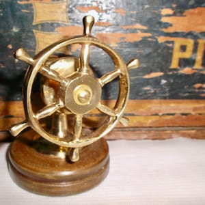 May include: A gold-toned brass ship's wheel with a wooden base. The wheel has five spokes and a small knob in the center.