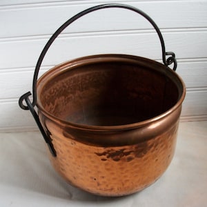 Puede incluir: Un caldero de cobre con textura martillada y un asa de metal negro. La olla tiene forma redondeada y una amplia abertura. El asa se arquea sobre la parte superior, sujeta a los lados. El caldero se utiliza probablemente para cocinar o con fines decorativos.