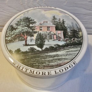 May include: A white ceramic jar with a lid featuring a hand-painted image of a house with a pink facade and snow-covered trees. The lid is inscribed with the words "Whitmore Lodge".