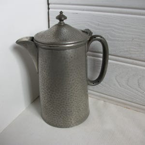 Può includere: Una brocca vintage in metallo martellato con coperchio e manico curvo. La brocca ha un beccuccio e un piccolo pomello sulla parte superiore del coperchio. Il metallo ha un aspetto screziato e strutturato. La brocca è alta circa 25 cm.