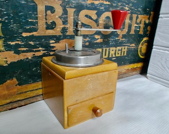 Puede incluir: Un molinillo de café vintage de madera con una tapa de metal y una manija de plástico roja. El molinillo tiene un cajón para guardar los granos de café.