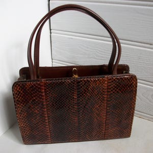 May include: A vintage brown handbag with a snakeskin pattern. The rectangular bag has two curved handles and a gold-colored clasp. The bag's exterior is a rich brown color with darker brown markings.