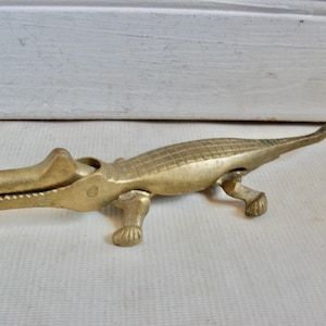 May include: A brass alligator-shaped nutcracker. The nutcracker is detailed with scales and has a hinged jaw. The alligator is resting on four legs and has a long tail. The nutcracker is a decorative and functional item.