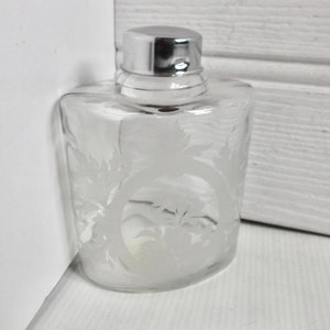 Könnte beinhalten: Klarer Parfümflakon aus Glas mit silberfarbenem Verschluss. Die Flasche hat eine abgerundete rechteckige Form und ist mit einem geätzten Blumendesign verziert. Die Flasche ist leer und steht auf einer weißen Oberfläche.