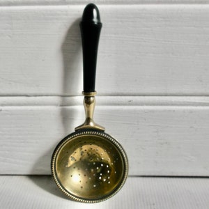 May include: A gold tea strainer with a black wooden handle. The strainer has a round, perforated bowl and a beaded rim.