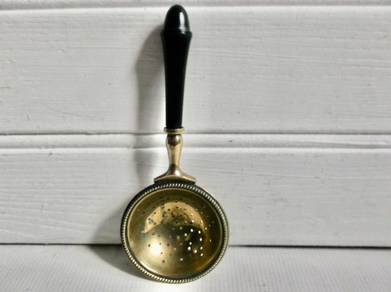 May include: A gold tea strainer with a black wooden handle. The strainer has a round, perforated bowl and a beaded rim.