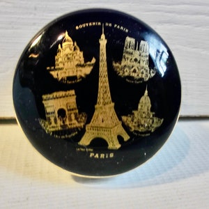 May include: A black oval trinket box with a gold trim and a gold design of famous Parisian landmarks, including the Eiffel Tower, Notre Dame Cathedral, the Arc de Triomphe, and Sacré-Cœur Basilica. The box reads "Souvenir de Paris" and "Paris".