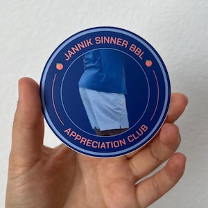 Puede incluir: Una pegatina redonda con un fondo azul oscuro y texto blanco que dice "JANNIK SINNER BBL APPRECIATION CLUB". La pegatina presenta una imagen parcial de una persona que lleva una camisa azul y pantalones cortos blancos. Hay ilustraciones de melocotones en la pegatina.
