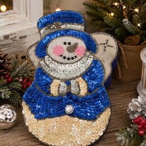 May include: A decorative snowman ornament crafted with blue, silver, and cream-colored sequins. The snowman features a blue top hat, a silver sequined scarf, and a face detailed with small beads, including pink cheeks and a dark nose.