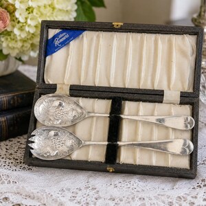 May include: A pair of silver-plated serving utensils, a spoon and a fork, featuring embossed fruit motifs on the bowls. They are presented in a dark rectangular hinged case with a cream-colored pleated satin lining and a blue label reading Rammers.