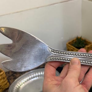 May include: A silver-toned metal serving utensil with a three-pronged, wide head and a handle featuring a decorative beaded border design. The utensil has a polished, reflective surface and a distinct, elongated shape for serving food items.