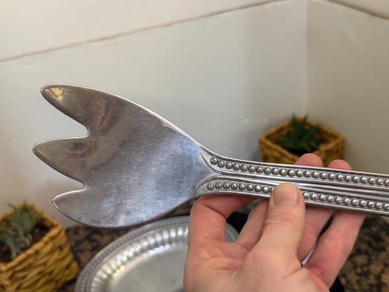 May include: A silver-toned metal serving utensil with a three-pronged, wide head and a handle featuring a decorative beaded border design. The utensil has a polished, reflective surface and a distinct, elongated shape for serving food items.