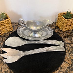 May include: A silver-toned metal gravy boat with a matching oval underplate, accompanied by a large serving spoon and a three-pronged serving fork. Each piece features a decorative beaded border along the edges and a smooth, polished finish.