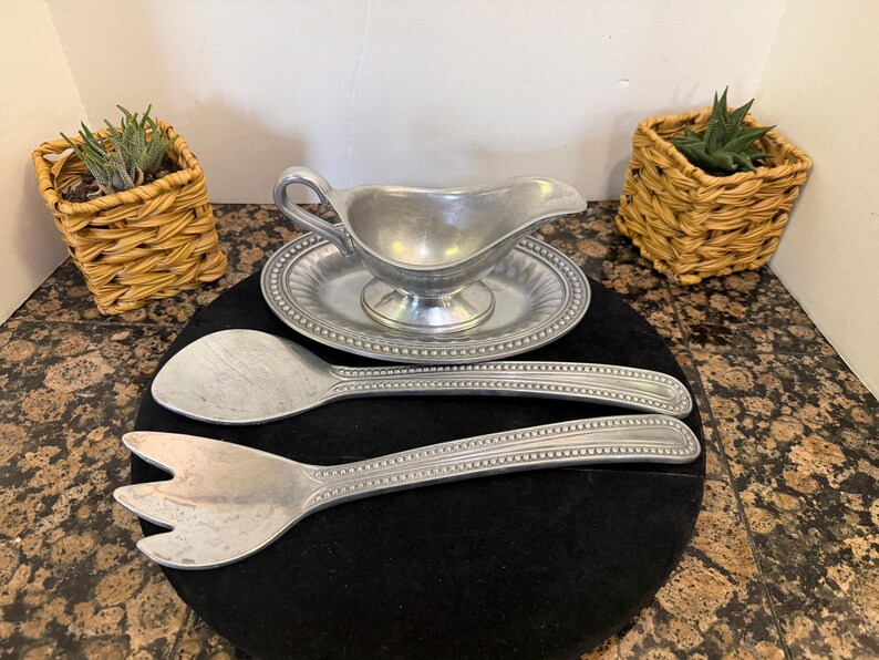 May include: A silver-toned metal gravy boat with a matching oval underplate, accompanied by a large serving spoon and a three-pronged serving fork. Each piece features a decorative beaded border along the edges and a smooth, polished finish.