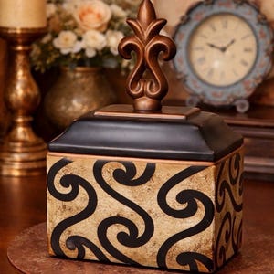 May include: A decorative box with a black top and a gold-colored body featuring a black swirl pattern. The box is topped with a bronze-colored finial. The box is sitting on a brown surface.