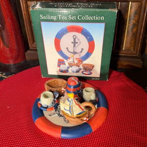 May include: A nautical-themed tea set collection featuring a teapot, cups, and bowls, all arranged on a lifesaver-shaped tray. The set includes a box with the words "Sailing Tea Set Collection" and a graphic of a life preserver and anchor.