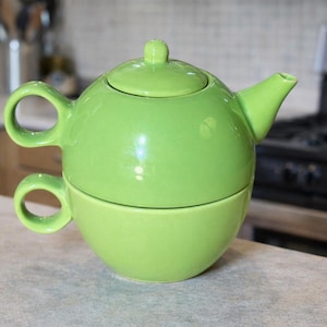 May include: A bright green ceramic teapot and teacup set. The teapot has a rounded shape with a lid and spout. The teacup is stacked beneath the teapot, with a handle on each piece. The set is on a countertop.