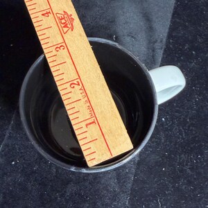 May include: A black coffee mug with a light grey handle. A wooden ruler is placed inside the mug, showing a measurement of approximately 7.6 cm. The ruler has red markings and the text "Made in U.S.A."