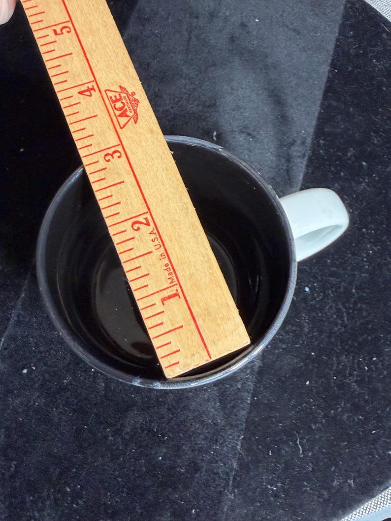 May include: A black coffee mug with a light grey handle. A wooden ruler is placed inside the mug, showing a measurement of approximately 7.6 cm. The ruler has red markings and the text "Made in U.S.A."