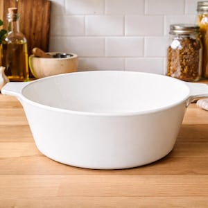 May include: A white, round, ceramic serving bowl with two handles. The bowl is empty and sits on a wooden countertop. Kitchen items, including herbs, garlic, and jars of food, are in the background.