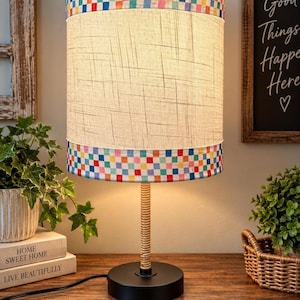 May include: A table lamp with a cylindrical shade featuring a beige linen-textured center band and multicolored checkered borders at the top and bottom. The lamp has a thin, wrapped stem and a solid black circular base with a black power cord.