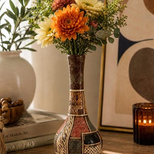 May include: A tall, slender ceramic vase with a narrow neck and a bulbous base. The surface features a patchwork of textured geometric patterns in shades of brown, cream, black, and terracotta, filled with a bouquet of orange, yellow, and green flowers.