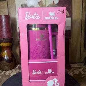 May include: A pink Stanley travel mug with a matching straw, packaged in a pink Barbie-branded box. The box features the Barbie logo, the Stanley logo, and a silhouette of Barbie's profile. The mug has the Stanley logo and the Barbie logo.