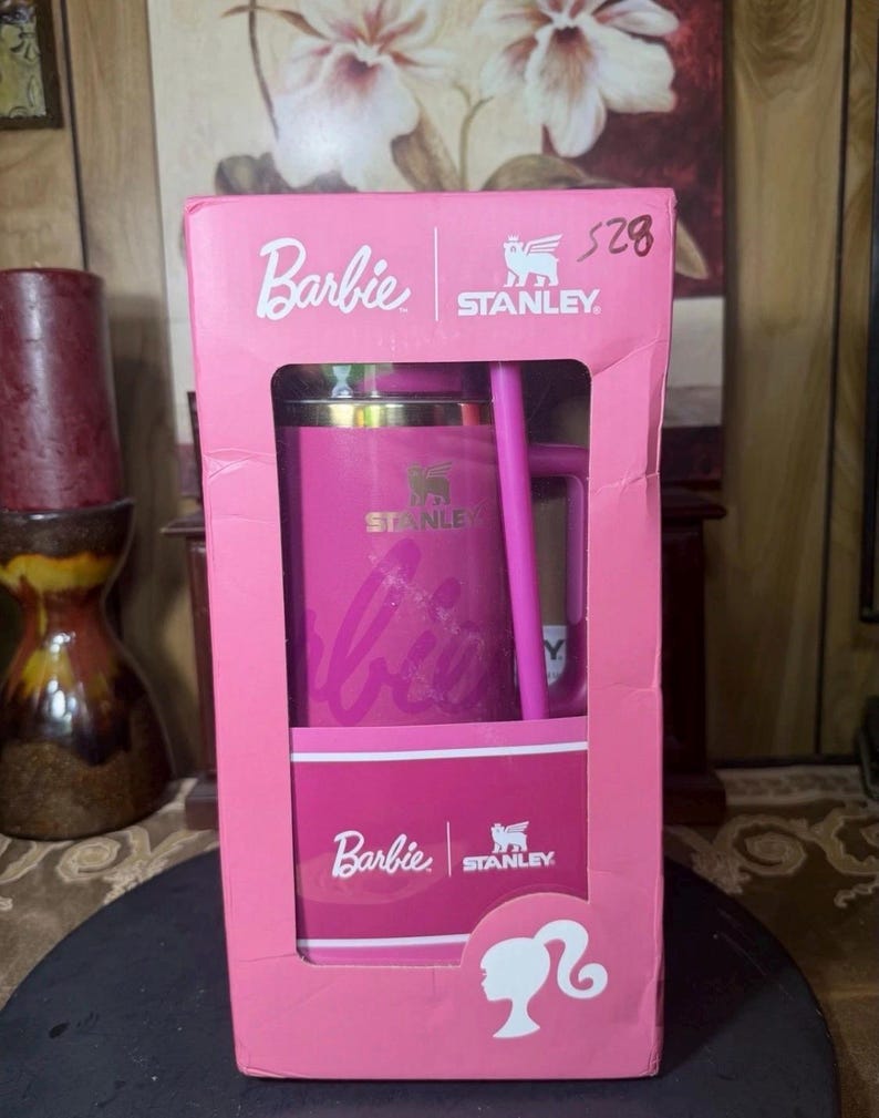 May include: A pink Stanley travel mug with a matching straw, packaged in a pink Barbie-branded box. The box features the Barbie logo, the Stanley logo, and a silhouette of Barbie's profile. The mug has the Stanley logo and the Barbie logo.