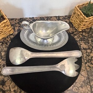 May include: A silver-toned metal gravy boat with an attached oval base featuring a beaded rim, accompanied by a matching large serving spoon and a three-pronged serving fork, each piece displaying a circular emblem on the handle.