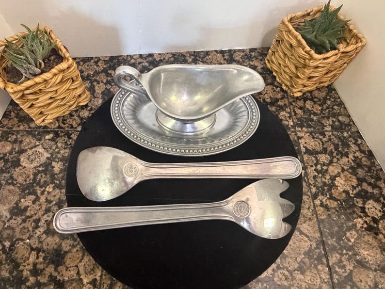 May include: A silver-toned metal gravy boat with an attached oval base featuring a beaded rim, accompanied by a matching large serving spoon and a three-pronged serving fork, each piece displaying a circular emblem on the handle.