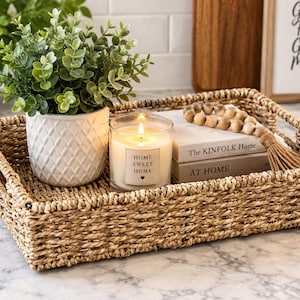 May include: A rectangular woven seagrass tray containing a white ceramic pot with a green plant, a glass candle labeled Home Sweet Home, two stacked books titled The Kinfolk Home and At Home, and a strand of wooden beads with a jute tassel.