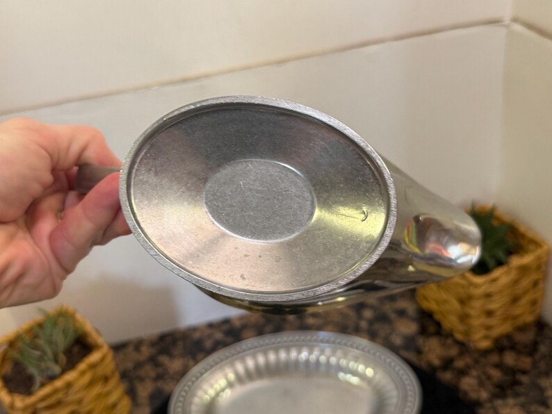 May include: The underside of a metallic gravy boat featuring a smooth, oval-shaped base with a slightly recessed center. The surface has a matte, brushed silver-tone finish with a visible circular texture pattern consistent with metal casting.