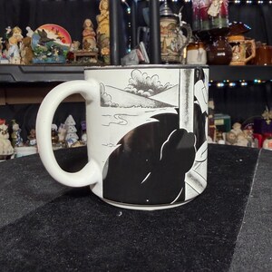 May include: White ceramic mug with a black and white comic strip design. The mug has a large handle and a wraparound illustration of a landscape and a silhouetted figure. The mug is set against a black surface.