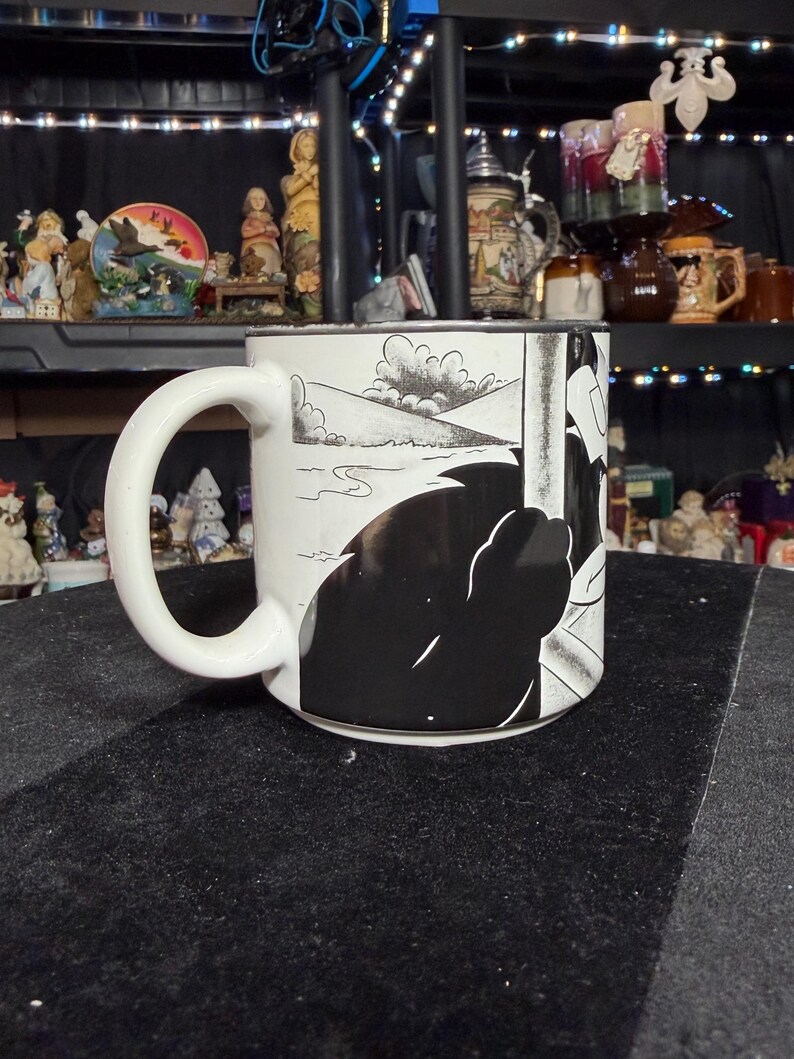 May include: White ceramic mug with a black and white comic strip design. The mug has a large handle and a wraparound illustration of a landscape and a silhouetted figure. The mug is set against a black surface.