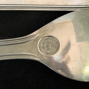 May include: A silver-toned metal serving fork with a smooth, reflective surface and a long handle. A circular stamp on the neck features the text RWP WILTON ARMETALE and the website address WWW.ARMETALE.COM, with a central logo containing the letters AWP.