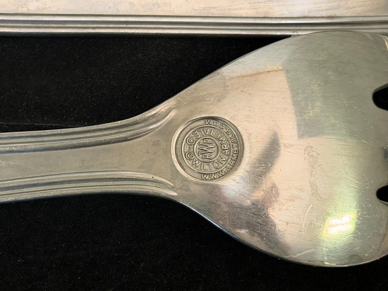 May include: A silver-toned metal serving fork with a smooth, reflective surface and a long handle. A circular stamp on the neck features the text RWP WILTON ARMETALE and the website address WWW.ARMETALE.COM, with a central logo containing the letters AWP.