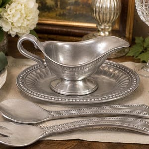 May include: A silver-toned metal gravy boat with a matching oval underplate, accompanied by a large serving spoon and a three-pronged serving fork. Each piece features a decorative beaded edge detail and a smooth, brushed metallic finish.